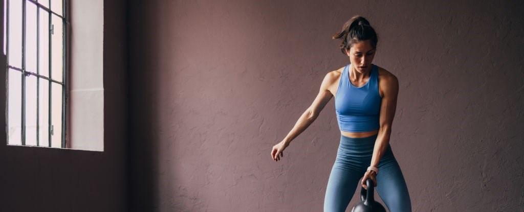 A fit woman in athletic wear lifting a kettlebell, concentrating on her workout in a muted gym environment.